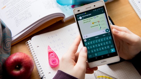 Eine Schülerin sitzt mit ihrem Handy und ihren Schulsachen an einem Schreibtisch und hat einen Chat geöffnet.