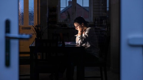 Eine Frau sitzt am Schreibtisch und schaut nachdenklich in ihren Laptop, während es draußen dunkel ist