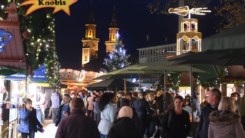 Zahlreiche Besucher gehen über den Weihnachtsmarkt in Ludwigshafen - beleuchtete Buden, eine Weihnachtspyramie und die Türme der Kirche St. Ludwig in der Innenstadt sind zu sehen.