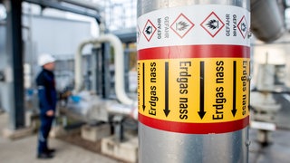 Rohre mit der Aufschrift "Erdgas nass" verlaufen an einer technischen Anlage zur Kühlung von Erdgas - im Hintergrund steht ein Arbeiter in blauer Arbeitskleidung mit weißem Helm und prüft die Anlage.