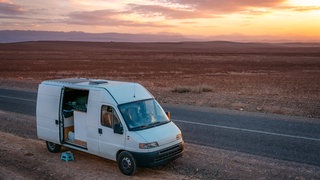 Ein Camper parkt in einer menschenleeren Umgebung am Straßenrand während die Sonne untergeht.