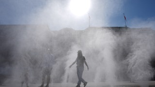 Passanten gehen durch einen Sprühnebel eines Wasserzerstäubers in der Innenstadt. Im Hintergrund sind durch den Nebel Häuser zu sehen. Dahinter blendet die Sonne.
