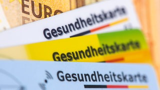 ILLUSTRATION: Verschiedene Gesundheitskarten von Krankenversicherungen liegen zusammen. Im Hintergrund wird eine 50 Euro-Banknote abgebildet.