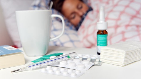 Grippesaison in Rheinland-Pfalz hat begonnen. Eine Frau liegt mit Grippe im Bett. Im Vordergrund sind Tabletten, Nasenspray und Taschentücher zu sehen.