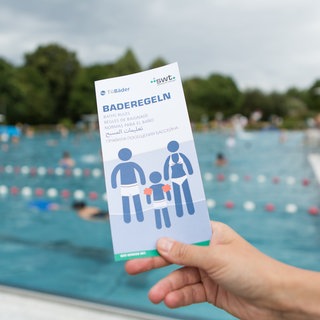 Becken im Freibad. Im Vordergrund eine Hand, die einen Flyer mit der Aufschrift "Baderegeln" hält. Darin steht, was erlaubt und was verboten ist.