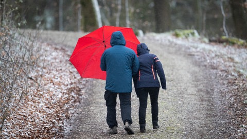 Spaziergänger mit einem roten Regenschirm auf einem schneebedeckten Wanderweg. Das Wetter in RLP bringt Schnee und Graupel