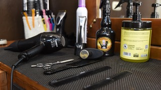 Scheren, Kämme und ein Fön liegen vor Haarwachs und einem Spiegel in einem Barbershop auf einem kleinen Tisch. 