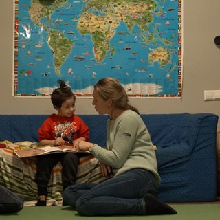Ein Kind sitzt in der Spielstube Stuttgart Weilimdorf auf einem Sofa und schaut sich gemeinsam mit einer Betreuerin ein Buch an.