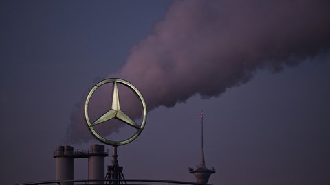 Der Mercedes Stern, das Logo des Fahrzeugherstellers Mercedes-Benz steht auf einem Gebäude einer Niederlassung, dahinter steigt eine Wolke aus einem Schornstein auf. Mercedes hat sich mit dem Gesamtbetriebsrat auf einen Sparpogramm geeinigt. 