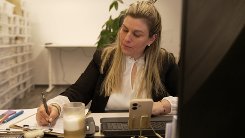 Eine Frau sitzt an einem Schreibtisch mit Smartphone und Tastatur und schreibt auf einen Zettel auf einem Klemmbrett. Anna-Maria Rock ist selbstständige Hochtzeitsplaner und hat drei Kinder. 