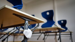 Symbolbild | Mundschutzmaske hängt an einem Schultisch in einem Klassenzimmer mit auf den Tischen aufgestellten Stühlen.