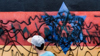 Es ist zu sehen, dass Unbekannte mit schwarzer Farbe einen Davidstern auf einer schwarz-rot-goldenen Wand durchgestrichen haben. Sie zerstörten damit ein Werk des Künstlers G. Schaefer an der East Side Gallery in Berlin. 