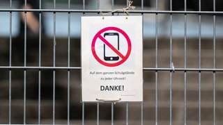 Ein Schild mit einem durchgestrichenen Smartphone und der Aufschrift "Auf dem ganzen Schulgelände - zu jeder Uhrzeit! DANKE!" hängt am Zugangstor zum Schulhof eines Gymnasiums und weist auf ein generelles Handyverbot hin.