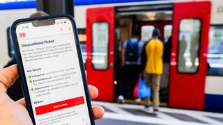 (Symbolbild) Eine Information zum Kauf des Deutschlandtickets ist auf dem Display eines Smartphones an einem Bahnhof zu sehen. 