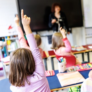 Schüler einer gemischten ersten und zweiten Klasse sitzen während des Unterrichts auf ihren Plätzen und heben die Hände. Mit Blick auf 1.440 unwissentlich unbesetzte Lehrerstellen in Baden-Württemberg gibt es noch viele offenen Fragen.