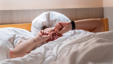 Ein junger Mann liegt im Bett und reibt sich beim Aufwachen müde die Augen, während er erschöpft und verschlafen in den neuen Tag startet.