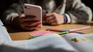 Ein Schulkind sitzt am Schreibtisch, bearbeitet Aufgaben mit einem Stift und hält dabei ein Smartphone in der Hand. (Symbolbild)