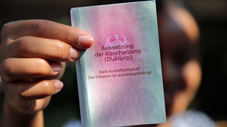 Eine junge Frau aus Afrika hält einen Ausweis für Flüchtlinge in der Hand mit dem Titel: "Aussetzung der Abschiebung (Duldung)".