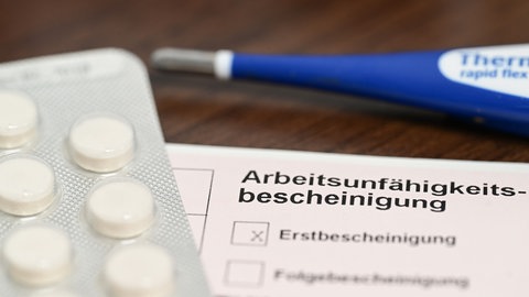 Ein Fieberthermometer, Medikamente und eine ärztliche Arbeitsunfähigkeitsbescheinigung (Krankmeldung) liegen auf einem Nachttisch.