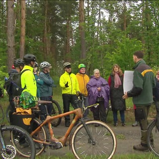 Eine Gruppe Radfahrer steht im Wald um einen Mann herum