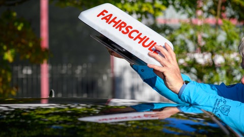 "Fahrschule" ist auf einem Schild zu lesen das ein Fahrlehrer auf das Dach seines Autos stellt.