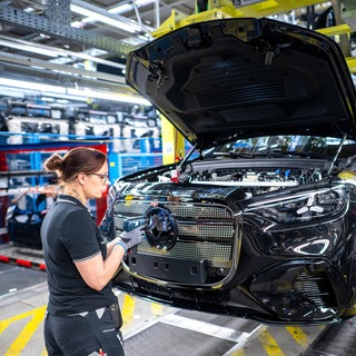 Beschäftigte arbeiten in einer Halle in einem Mercedes-Benz Werk an einem Fließband an Fahrzeugen vom Typ GLC.