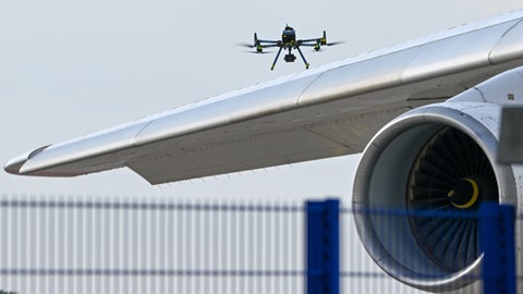 Symbolbild: Eine Drohne schwebt über dem Flügel eines Flugzeugs. Nach dem Fund einer Drohne mit Sprengstoff am Flughafen LeipzigHalle verlässt man sich an Flughäfen in Baden-Württemberg auf die bereits bestehenden Sicherheitsmaßnahmenac