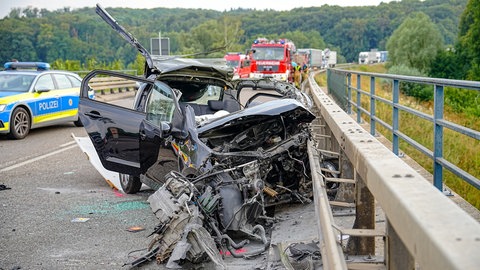Ein zerstörtes Fahrzeug steht auf an der Unfallstelle, daneben ein Polizeiauto und ein Feuerwehr-Wagen.
