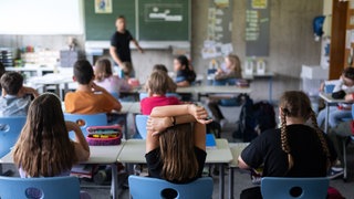 Schülerinnen und Schüler sitzen in einem Klassenzimmer. Im Hintergrund ist verschwommen ein Lehrer zu sehen, der vor der Tafel steht.