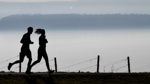Jogger laufen auf einem Wanderweg in Richtung Meersburg, während im Hintergrund die Stadt Konstanz am Bodensee im Dunst zu sehen ist. 