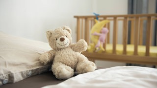 Auf einem Bett sitzt ein beiger Teddybär. Im Hintergrund steht ein Kinderbett.