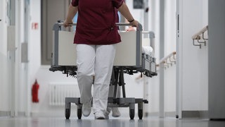 Eine Krankenpflegerin und ein Krankenpfleger schieben ein Krankenbett durch den Gang einer Station eines Krankenhauses.