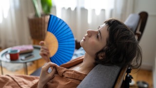 Eine Frau sitzt erschöpft auf dem Sofa und fächert sich Luft zu.