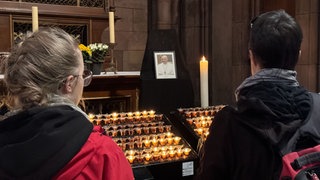 Zwei Menschen stehen vor vielen Kerzen. Über ihnen hängt ein Fotot des verstorbenen Papstes Franziskus