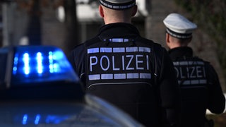 Das Logo der Polizei auf der Dienstkleidung von Polizisten die vor einem Blaulicht stehen. 