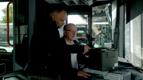 Zwei Männer stehen am Eingangsbereich eines Linienbusses und setzen sich mit der Kasse neben der Fahrerkabine auseinander.