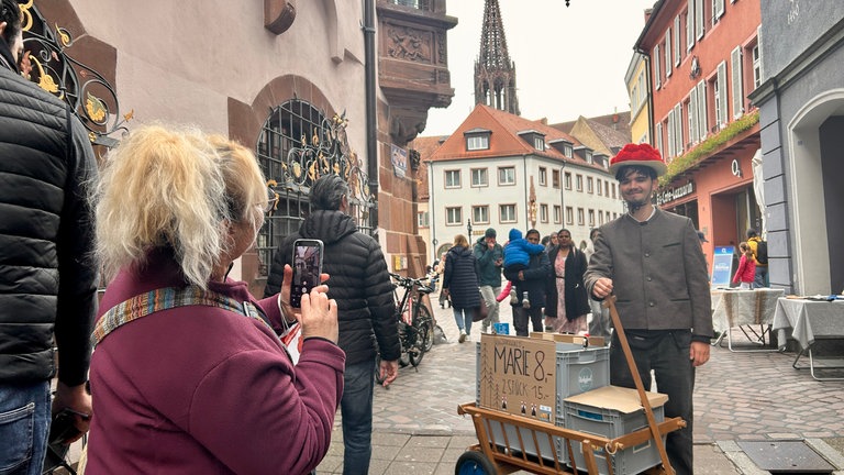 Ein junger Mann läuft mit Bollenhut und Bollerwagen durch Freiburg, um die Playmobil-Figur "Schwarzwald Marie" auf der Sztaße zu verkaufen.