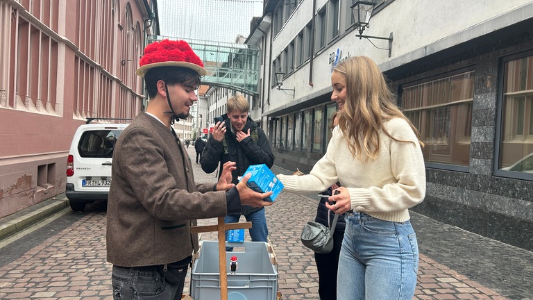Ein junger Mann läuft mit Bollenhut und Bollerwagen durch Freiburg, um die Playmobil-Figur "Schwarzwald Marie" auf der Sztaße zu verkaufen.