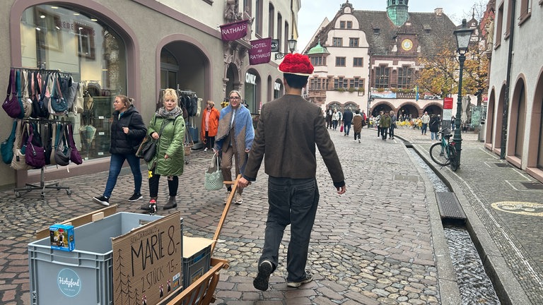 Ein junger Mann läuft mit Bollenhut und Bollerwagen durch Freiburg, um die Playmobil-Figur "Schwarzwald Marie" auf der Sztaße zu verkaufen.