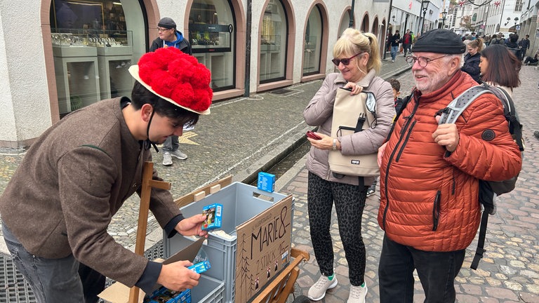 Ein junger Mann läuft mit Bollenhut und Bollerwagen durch Freiburg, um die Playmobil-Figur "Schwarzwald Marie" auf der Sztaße zu verkaufen.