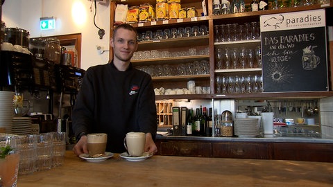 Ein junger Mann mit kurzen, blonden Haaren steht hinter einer Theke. Vor ihm stehen zwei Heißgetränke zum Servieren bereit.