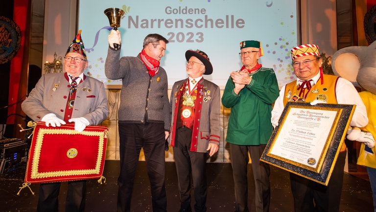 Markus Söder mit goldener Narrenschelle in der Hand, Wienfried Kreitschmann, der applaudiert, und närrische Vertreter.