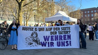 Zwei Demonstrierende halten ein Banner "Justiz & Uni auf der Seite der Täter? Wehren wir uns!"
