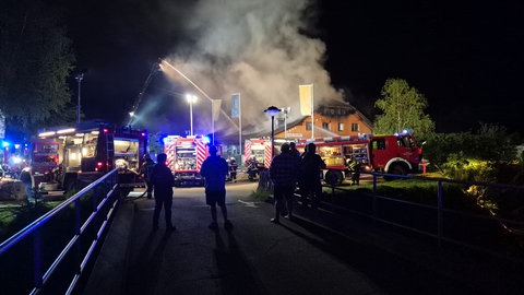 Im Vordergrund stehen einige Menschen im Schatten, im Hintergrund sieht man, wie die Feuerwehr versucht das Hauptgebäude des Campingplatz Bankenhofen zu löschen.