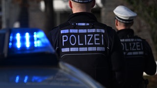 Das Logo der Polizei auf der Dienstkleidung von Polizisten die vor einem Blaulicht stehen (gestellte Szene).