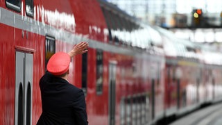 Ein Bahnmitarbeiter gibt an einem Bahnsteig ein Handsignal zur Weiterfahrt. 