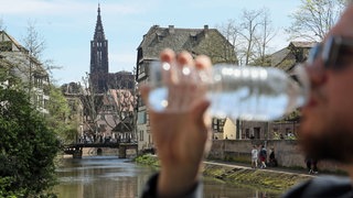 Symbolbild: Ein Mann trinkt einen Schluck Wasser aus einer Flasche. Im Hintergrund ist die Kathedrale von Straßburg zu sehen.