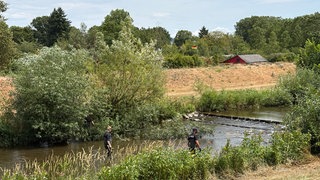 Zwei Männer mit Cappy  und schwarzen Tshirts und der Aufschrift "Polizei" steigen in einen kleinen Fluss.