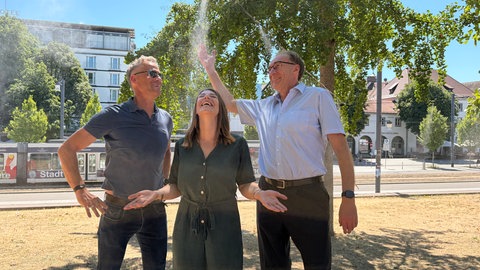 Frank Ueckermann, Leiter des Garten- und Tiefbauamts, die Projektverantwortliche für Grünoasen, Kathrin Sehl und Freiburgs Baubürgermeister Martin Haag lassen sich von der Vernebleranlage mit Wassertröpchen besprühen. 