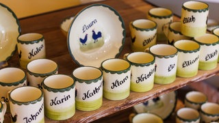 Tassen und ein Teller mit aufgemalten Vornamen und dem "Hahn und Henne"-Motiv stehen auf einem Holzbrett.